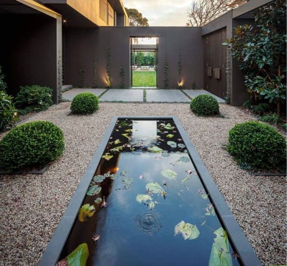 A simple rock garden with a minimalist ambiance featuring four dwarf evergreens, white pebble flooring, and a long koi pond.