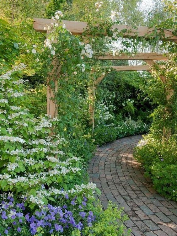 A garden pathway with a cottage vibe due to its brick flooring and wooden posts designed with creeping flowering plants. 